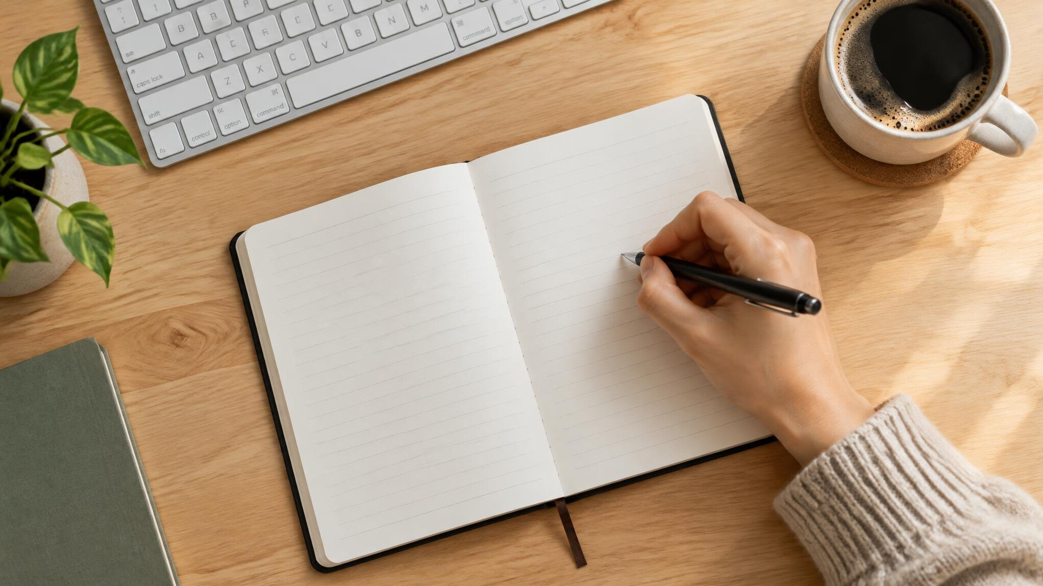 Hand writing in an open notebook on a desk next to a keyboard